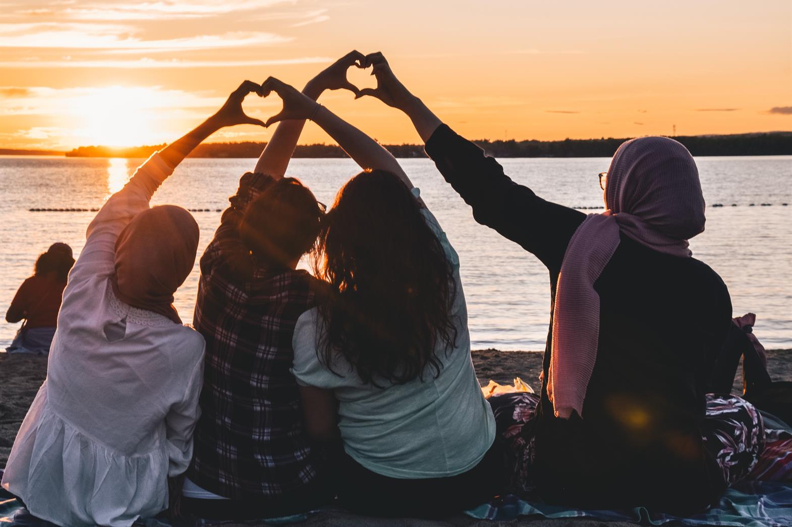 four people sitting on shore forming hearts with their hands during sunset