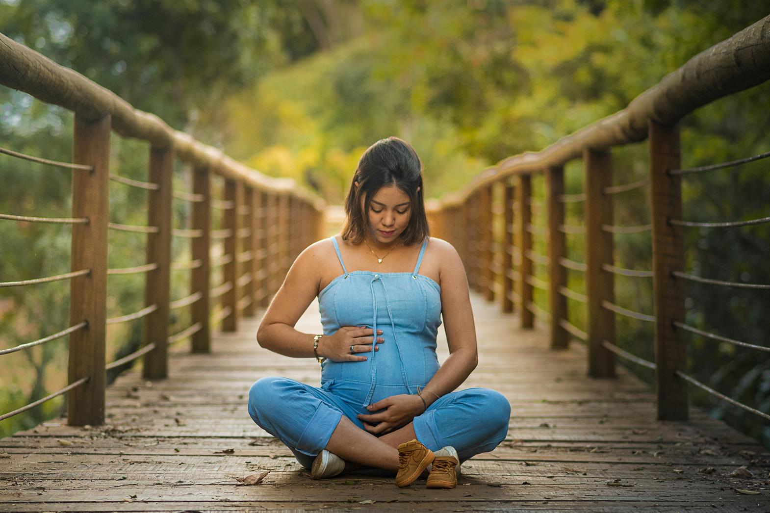Une femme enceinte assise en tailleur sur un pont en bois dans une forêt