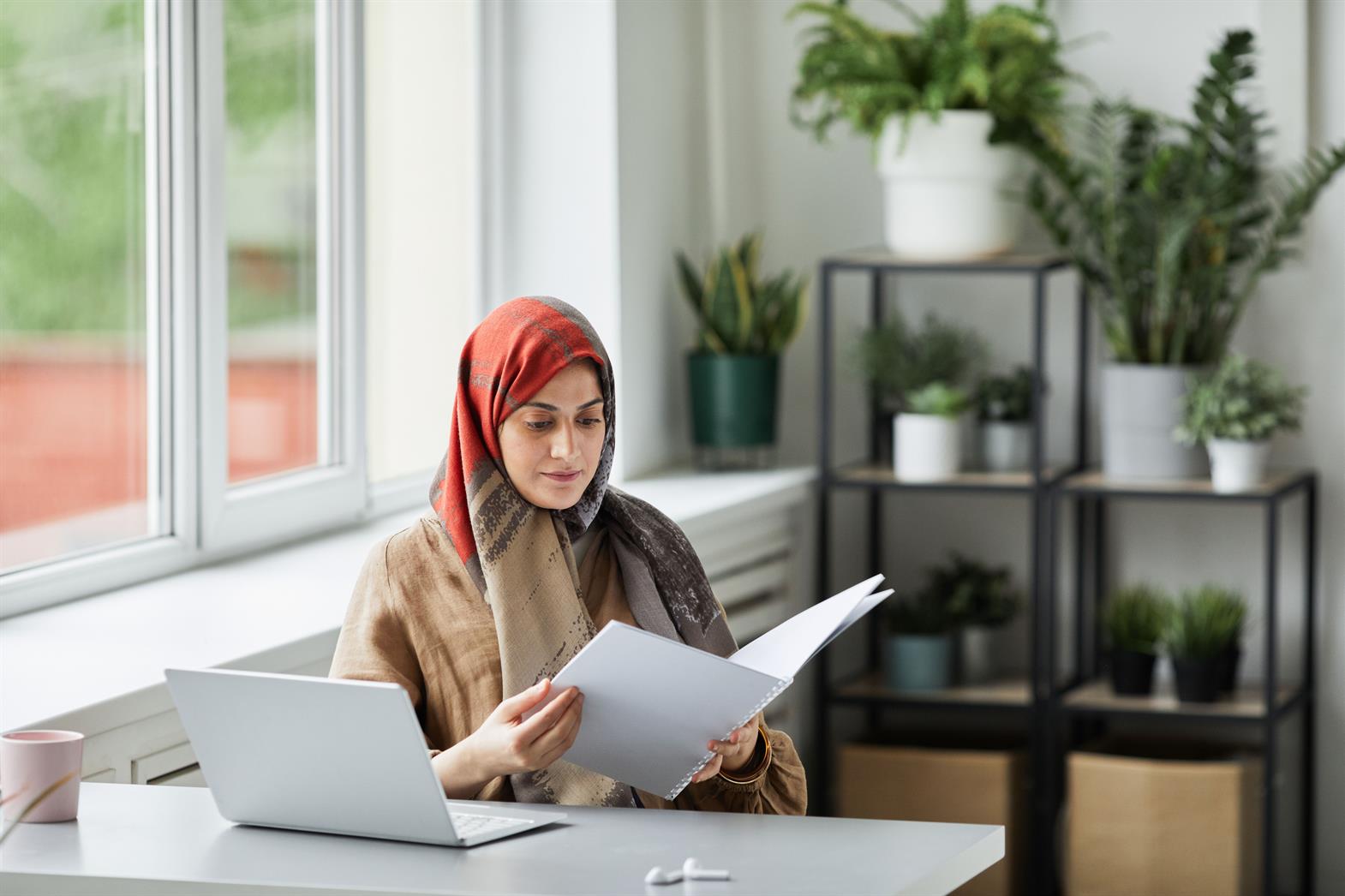A woman reviewing paperwork at desk with laptop and plants