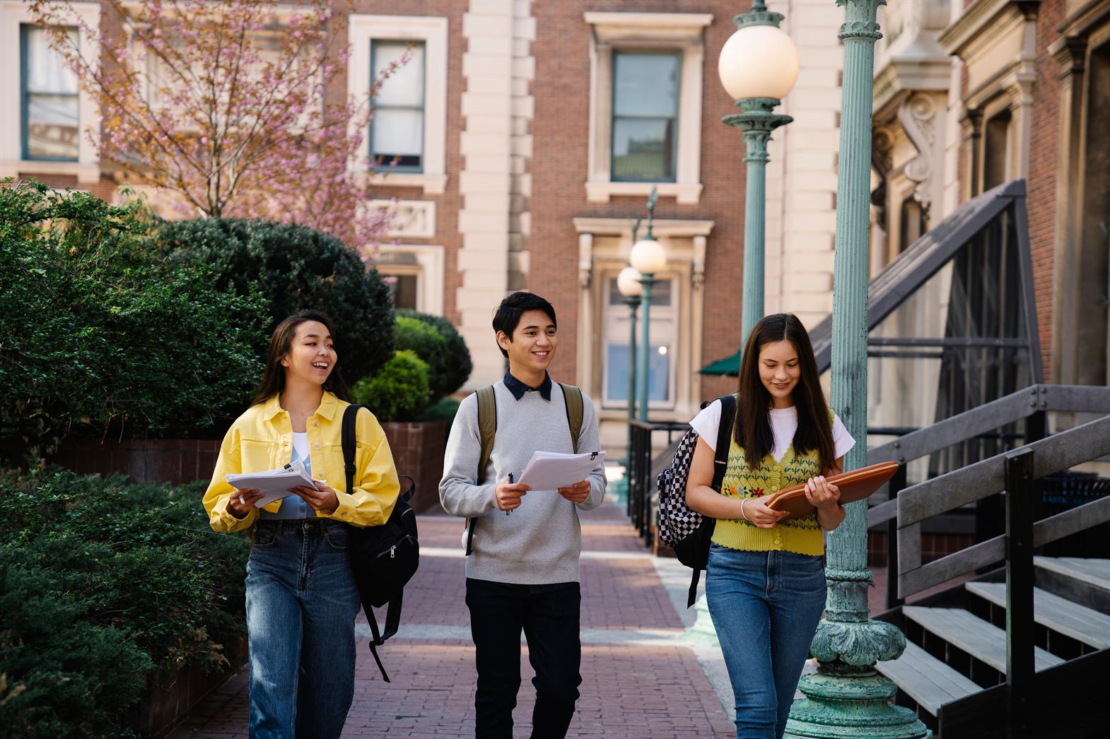 Three young adults walking and talking on a sunny campus day, holding notebooks and backpacks